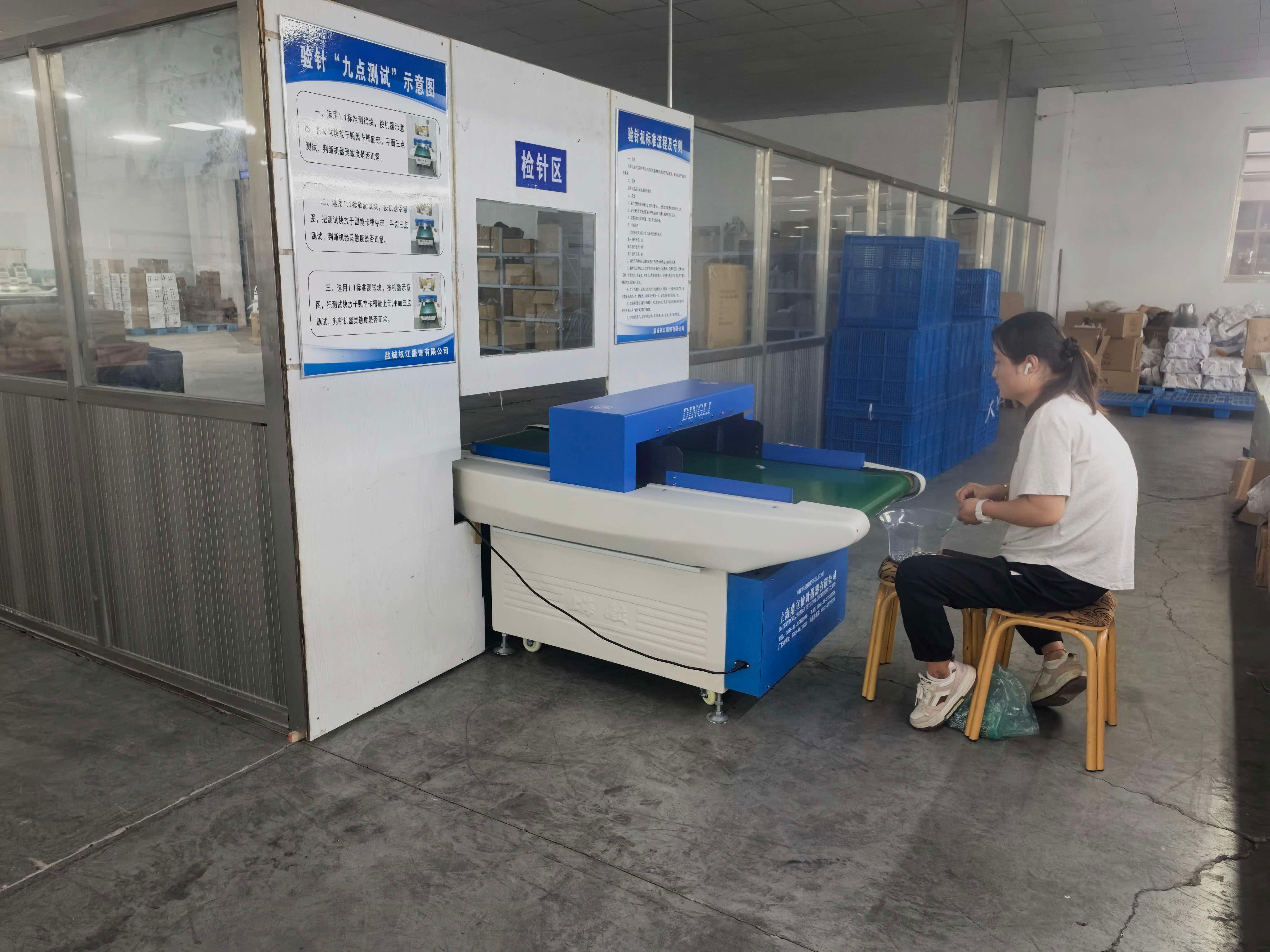 Inspection area with a conveyor needle detector, posted procedure sheets and a worker seated beside it